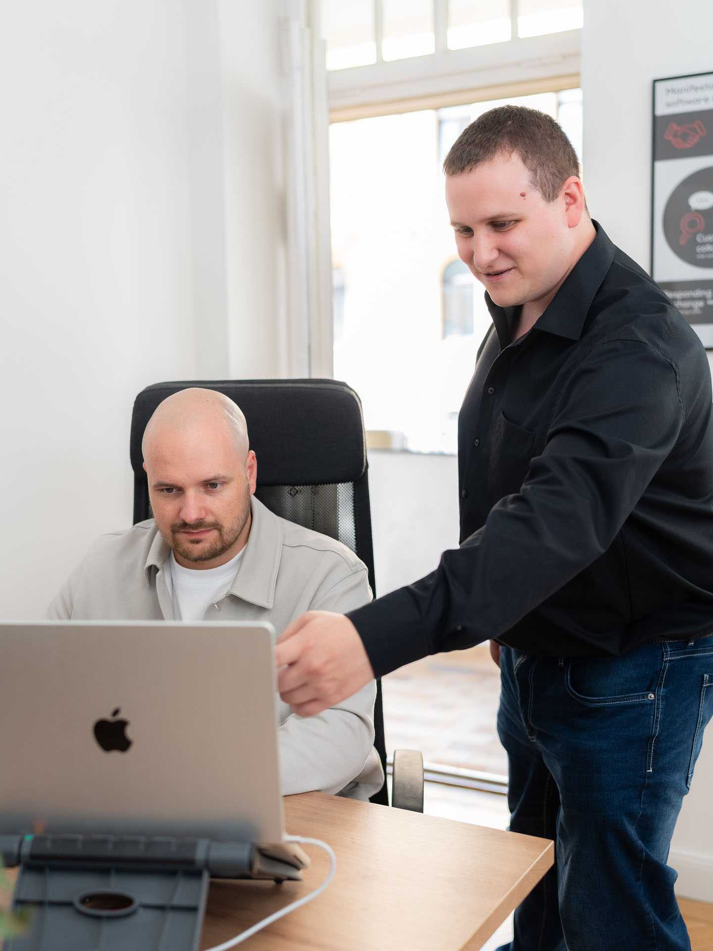 One employee is sitting in front of a laptop while another stands beside them, pointing at the screen.