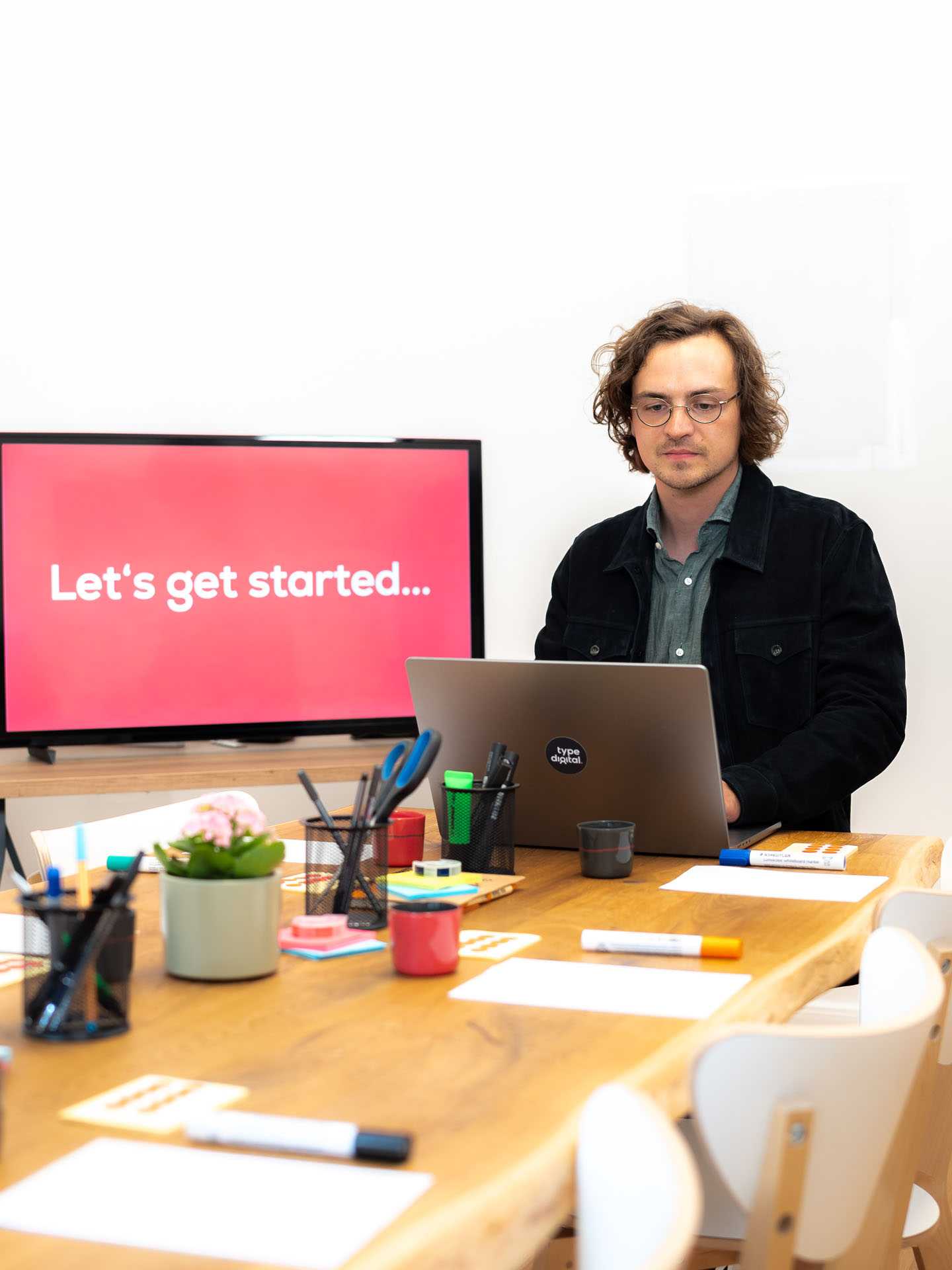 A man is sitting at the end of the table, looking at his laptop in front of him. There are workshop materials on the table. In the background, a television displays the phrase 'Let's get started...'